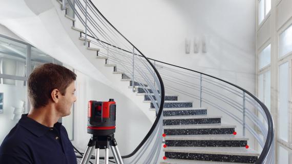Homme alignant un laser multiligne rouge-noir Leica sur un escalier en colimaçon dans un intérieur moderne blanc, points laser rouges visibles sur les marches