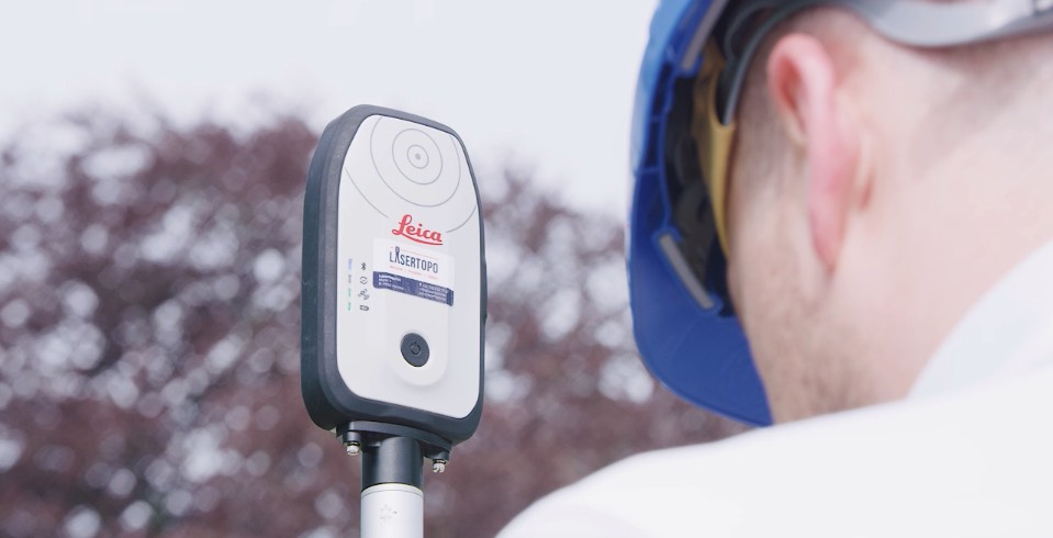 Gros plan d'une antenne GNSS Leica FLX100 avec autocollant Lasertopo sur une canne de mesure, utilisée par un géomètre avec casque bleu et lunettes de soleil